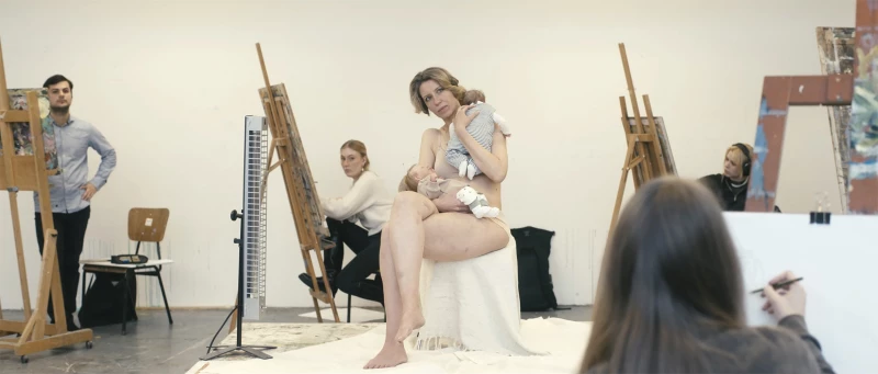Filmstill "Reproduktion". Ein Aktmodell sitzt mit zwei Babies im Arm inmitten von Studenten